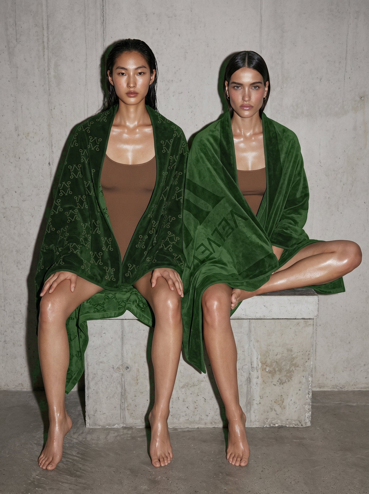 Two women wrapped in VELVET Undergrowth Monogram towels on a concrete bench