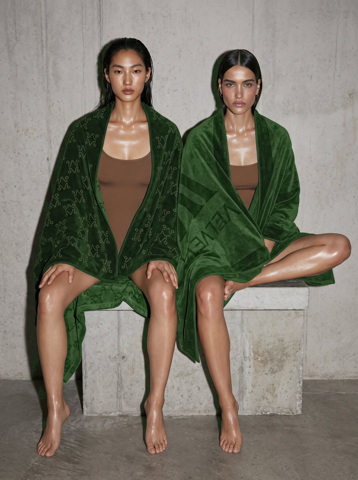Two women wrapped in VELVET Undergrowth Monogram towels on a concrete bench